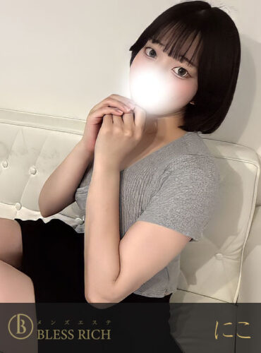 Woman with short dark hair sits on a white tufted sofa in a gray top and black skirt, hands near her face; logo watermark at bottom.