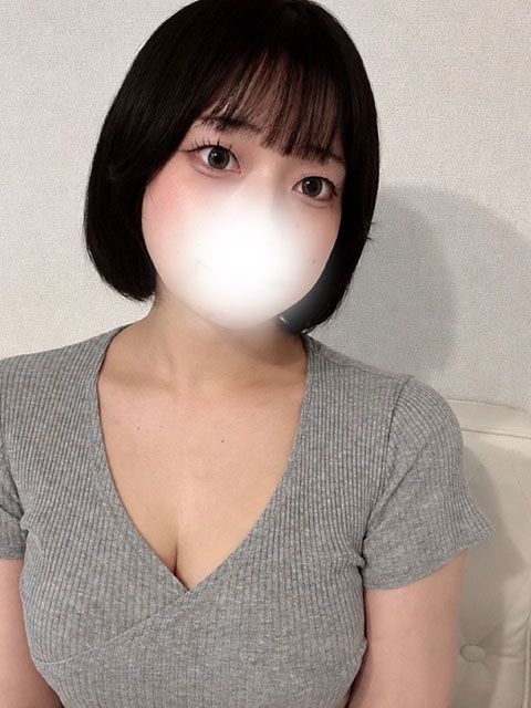 Woman with short black hair wearing a gray knit V-neck top, sitting on a light sofa; lower face obscured by a bright glare.